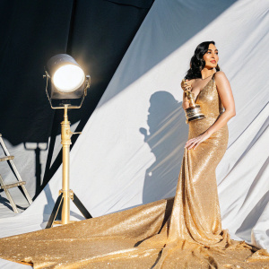 Gal Gadot at a glitzy awards show. Wearing a shimmering gold dress with a long train. Dark backdrop with spotlight