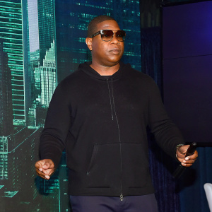 Ja Rule, wearing a black hoodie and sunglasses, at a tech conference. Modern cityscape backdrop.