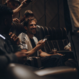 Leon Goretzka, wearing black and white jersey, is seated in a VIP lounge. Surrounded by fans, he poses for autographs and photos.