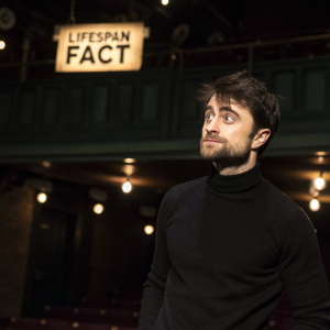Daniel Radcliffe, with scruffy dark hair and a beard, wears a black turtleneck. Standing in front of a dimly lit stage with 'LIFESPAN FACT' sign. An intimate theater setting.