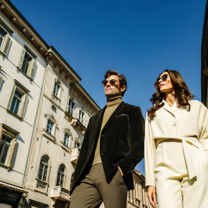 Jon Kortajarena, in sunglasses and a dark blazer, and Eugenia Silva, in a beige outfit, stand in front of an ornate European building. Bright sunny day with clear blue sky. Urban street scene.