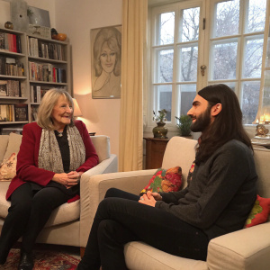 Alice Schwarzer and Conchita Wurst in a cozy living room setting, dressed in casual attire. A candid moment captured during an interview.