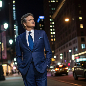 Colin Firth, dressed in a tailored blue suit with a matching blue tie and a crisp white shirt. Standing on a bustling city street at night.