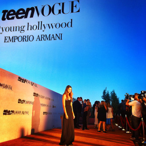 Kaia Gerber at the Teen Vogue Young Hollywood issue launch party. Wearing a black dress, standing against a white backdrop with Teen Vogue and Emporio Armani logos. Surrounded by guests and photographers.
