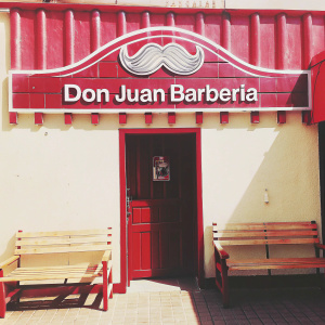 Photograph of Don Juan Barberia storefront. Vibrant logo featuring a stylized mustache and Spanish text. Bright yellow walls and wooden benches outside