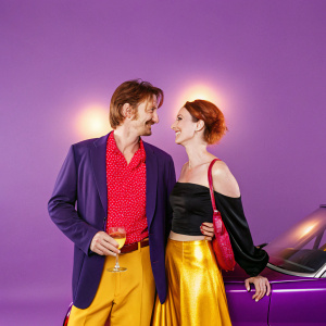 Sean Penn and Anna Bingemann at the film festival premiere. Sean wears a navy blazer with a red patterned shirt. Anna is in a black top and gold skirt. They share a friendly smile in front of a luxury car.