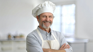 Caucasian middle aged male chef in a chef's hat with arms crossed wears apron standing in restaurant kitchen and smiling