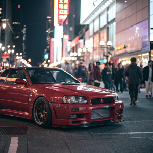 A shiny red Nissan Skyline R34 GTR parked in a busy downtown area. Bright lights and bustling crowd in the background.