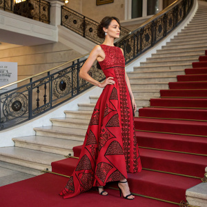 Olga Kurylenko at a film festival, standing in front of a grand staircase. Wearing a striking red dress with intricate patterns and high heels.