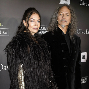 Lani Hammett and Kirk Hammett at the Pre-GRAMMY Gala. She is dressed in a black fluffy feathered outfit. He has long curly grey hair. They stand against a black backdrop with sponsor logos visible.