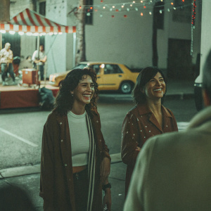 Nina Dobrev and Jessica Szohr, both smiling, at a lively street festival. A vibrant yellow taxi is in the background.