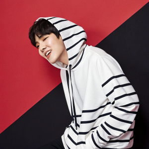 Suga from BTS in a casual photo shoot. Wearing a white and black striped hoodie. Dark, short hair styled neatly. Bold red and black background. Fun and relaxed pose.
