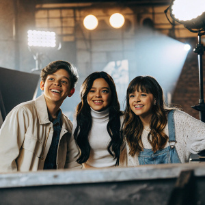 Actors from the show 'Stuck in the Middle', Isaac Presley, Jenna Ortega, and Lauren Pritchard, smiling for the camera. Set of a television studio, bright lights, colorful backdrop.