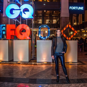 Ashley Greene at the GQ Super Bowl bash. She's in a black leather jacket and grey top. Behind her, the GQ and Fortune logos stand out against a white backdrop. Polished marble floor.