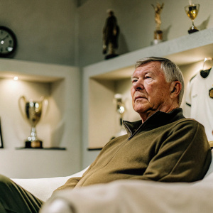 Sir Alex Ferguson, in a dimly lit interview room, surrounded by sports memorabilia. Solemn expression, deep-set eyes, sharp jawline. Emphasizing his leadership and wisdom.