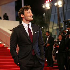 Sam Claflin attending the Cannes Film Festival premiere. Wears tailored black suit and maroon tie, styled short haircut.