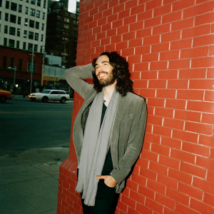 Russell Brand, bearded and smiling, in a gray scarf. Against a backdrop of busy city street.