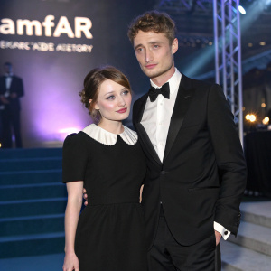 Emily Browning and Max Irons at the amfAR Gala. She's in a classic black dress with white collar, he's in a tuxedo. They pose against a backdrop with amfAR logo and large stage.
