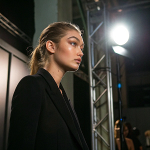 Gigi Hadid, a young model, in a black blazer. Standing in a backstage area at a fashion show. Spotlight illuminating her face