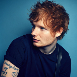 Close-up of Ed Sheeran. Red hair, blue background. Tattoo visible on his arm. Signature Ed Sheeran