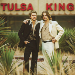 Garrett Hedlund and Jay Will attending the grand premiere of 'Tulsa King'. Dressed in black suit and patterned shirt respectively, they pose confidently in front of a dramatic red backdrop with bold lettering.