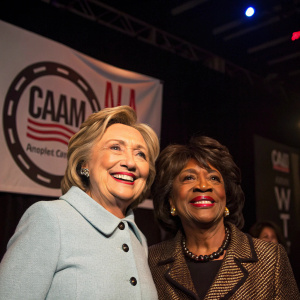 Hillary Clinton and Maxine Waters smiling on a dimly lit stage. Banner with CAAM logo behind them.