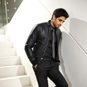 Sidharth Malhotra, in a sleek black leather jacket paired with a button-up shirt, tie, and dark slacks, poses against a bright white backdrop at a high-profile Bollywood premiere.