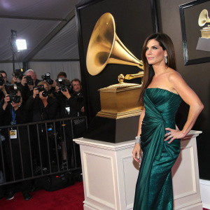 Sandra Bullock in a stunning emerald green dress, at the Grammy Awards. She's posing confidently, golden grammy statue beside her, paparazzi flashing in the background.