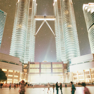 The Petronas Twin Towers in the heart of Kuala Lumpur's bustling cityscape. Neon lights and vibrant crowds animate the area. The towers stand tall against a starry night sky, their gleaming surfaces reflecting the city's vibrant energy.