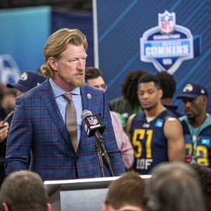 Les Snead, GM of the LA Rams, speaking at the NFL Combine. Sport coat and tie, professional demeanor. Athletes and reporters in background.