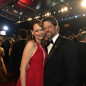Melinda Clarke and Ernie Mirich at a glamorous awards ceremony. She wears a flowing red evening gown, he is in a black tuxedo. They are hugging. Crowded indoor setting with spotlights.
