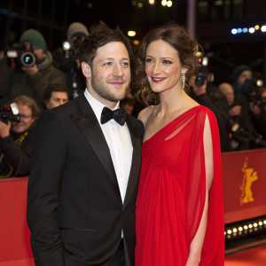 Matt Cardle and Sarah Robinson attending the Berlin Film Festival premiere. Both dressed in formal attire, Matt in a black tuxedo, Sarah in a flowing red dress. Crowds and flashing cameras in background.
