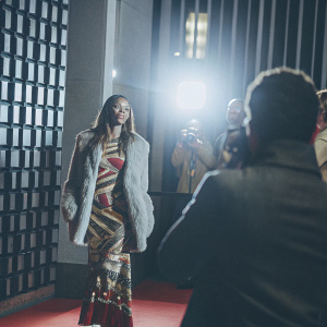 Fashion model at Black Panther: Wakanda Forever premiere. Red carpet event, bright lights and photographers. Wears colorful dress and furry jacket.