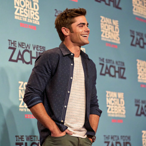 Zac Efron at the press junket. Dressed in casual attire, smiling brightly against a backdrop filled with promotional text.