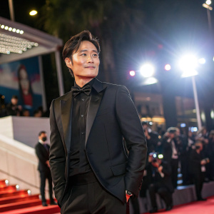Lee Byung-Hun, South Korean actor, dressed in a sleek black suit. Posing at Cannes Film Festival. Bright lights and bustling crowd in background.