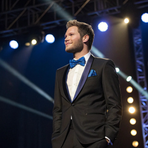 Jack Reynor in a sleek black tuxedo, with a blue bow tie. Standing solo on a stage under bright lights.
