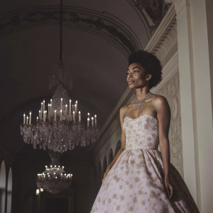 Yara Shahidi at the MET Gala. Pink floral dress with intricate beadwork and gold necklace. The venue is lavishly decorated with crystal chandeliers and opulent backdrops.