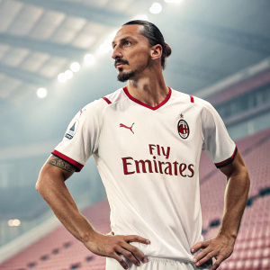 Zlatan Ibrahimovic, wearing a white and red AC Milan jersey with the Fly Emirates logo, posing confidently for a promotional photo shoot. Indoor stadium background with blurred rows of seats