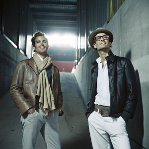 Spencer Matthews and Oliver Proudlock photographed at a Red Bull sponsored event. Spencer, left, in a brown leather jacket and tan scarf. Oliver, right, in black jacket, white shirt, and stylish hat.