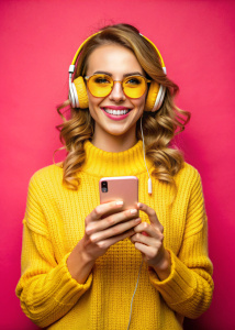 девушка блогер, на шее наушники, желтый свитер, улыбается, в руках смартфон, вид по пояс, темно-оранжевый однотонный фон, розовые очки