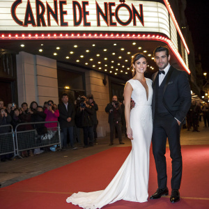 Blanca Suarez and Mario Casas attending the glamorous movie premiere of 'Carne de Neon'. Blanca is adorned in a stunning white floor-length gown, while Mario exudes elegance in a sleek black tuxedo. They stand beside a large marquee sign for the film premiere.