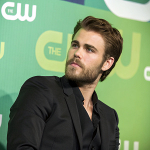 Paul Wesley in a formal black suit, posing at a press conference. The backdrop is a bright green with the CW Network logo. He has a serious expression and is well-groomed with a beard.