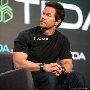 Close-up of Mark Wahlberg, actor and producer, at tech launch event. Black t-shirt, 'TICDA' logo, watch on wrist. Short beard, serious look. Brand logo backdrop