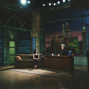 Rose McGowan on 'The Tonight Show with Jay Leno', Episode 3343. Rose seated on brown leather couch, Jay Leno standing at desk. Studio with colorful cityscape backdrop, warm lighting, lively atmosphere.