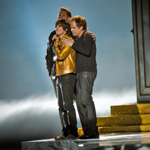 Kiefer Sutherland, Ben Stiller, and Zac Efron center stage at the 2009 MTV Movie Awards. Kiefer Sutherland in an embrace, Ben Stiller focused expression. Dramatic blue lighting backdrop.