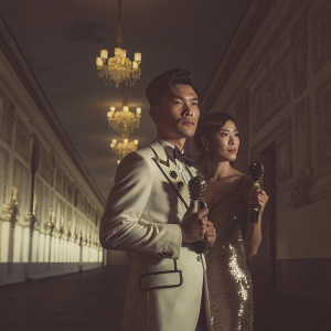 Ruco Chan and Nancy Wu at the Dragon Award Ceremony. Ruco in a white suit with black accents. Nancy in a shimmering black gown. They stand with their trophies in a grand hall with gold detailing.