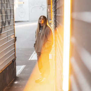 Avril Lavigne, in casual attire, standing alone in a vibrant, neon-lit alleyway. Graffiti and street art fill the background.