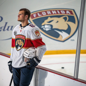 Aleksander Barkov, the NHL star, standing confidently with his helmet in hand. Ice rink in background, team Florida Panthers logo visible