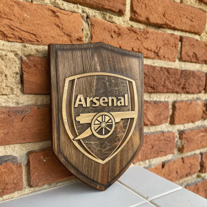 Vintage Arsenal logo design. Engraved on wooden plaque. Displayed against a rustic brick wall background.