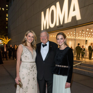 Susan Rockefeller, David Rockefeller Sr., and Ariana Rockefeller attending a gala at the Museum of Modern Art. They pose for photographers in front of a large, illuminated backdrop featuring the MOMA logo.